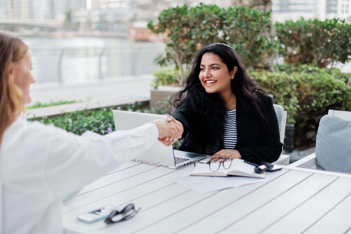 Two professional women shake hands across a white table at an outdoor cafe during a successful business meeting