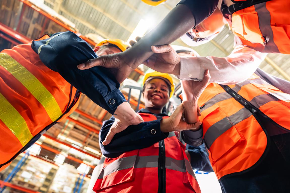 A diverse team of construction workers in high-visibility vests and hard hats link their arms in a circle, smiling up at the camera in a show of teamwork.