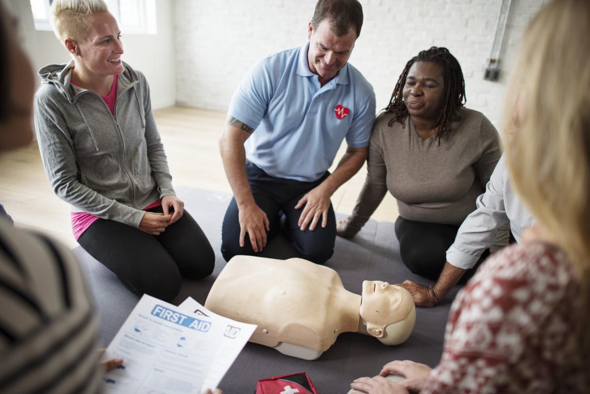 A group of employees in a bright room participate in a first aid training session, gathered around a CPR dummy as an instructor demonstrates.