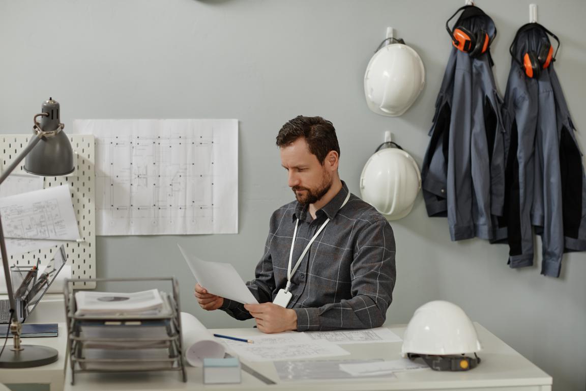 A male site manager wearing an ID card lanyard sits at his office desk, carefully reviewing safety documents. In the background, hard hats and work jackets hang on the wall.