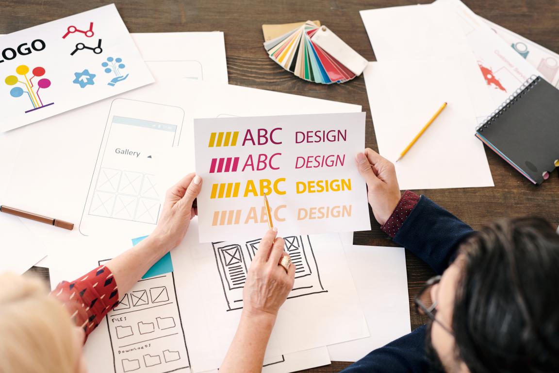 Two designers collaborate over a desk covered in sketches, one holding up a sheet with different font and colour options for a brand design