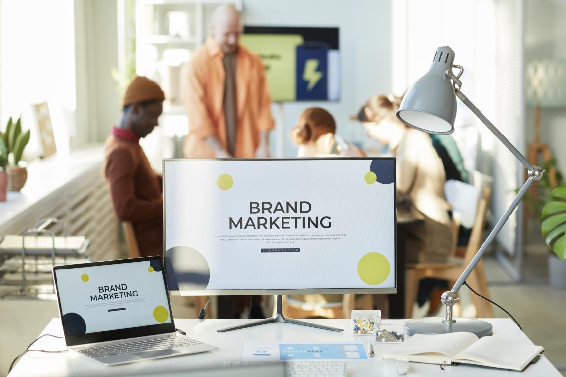 A computer monitor and laptop in a modern office both display a 'BRAND MARKETING' presentation slide, with colleagues working in the background.