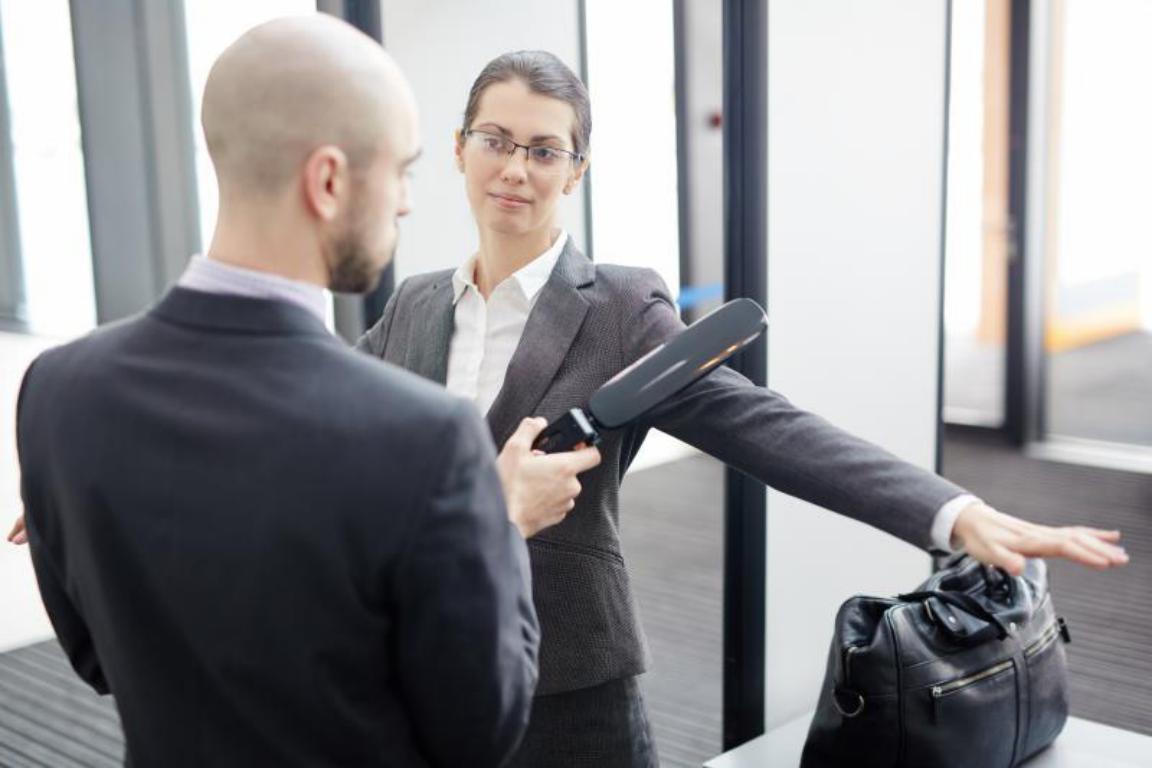 A UK airport security officer performs a standard secondary screening with a hand-held metal detector on a passenger with a medical implant.
