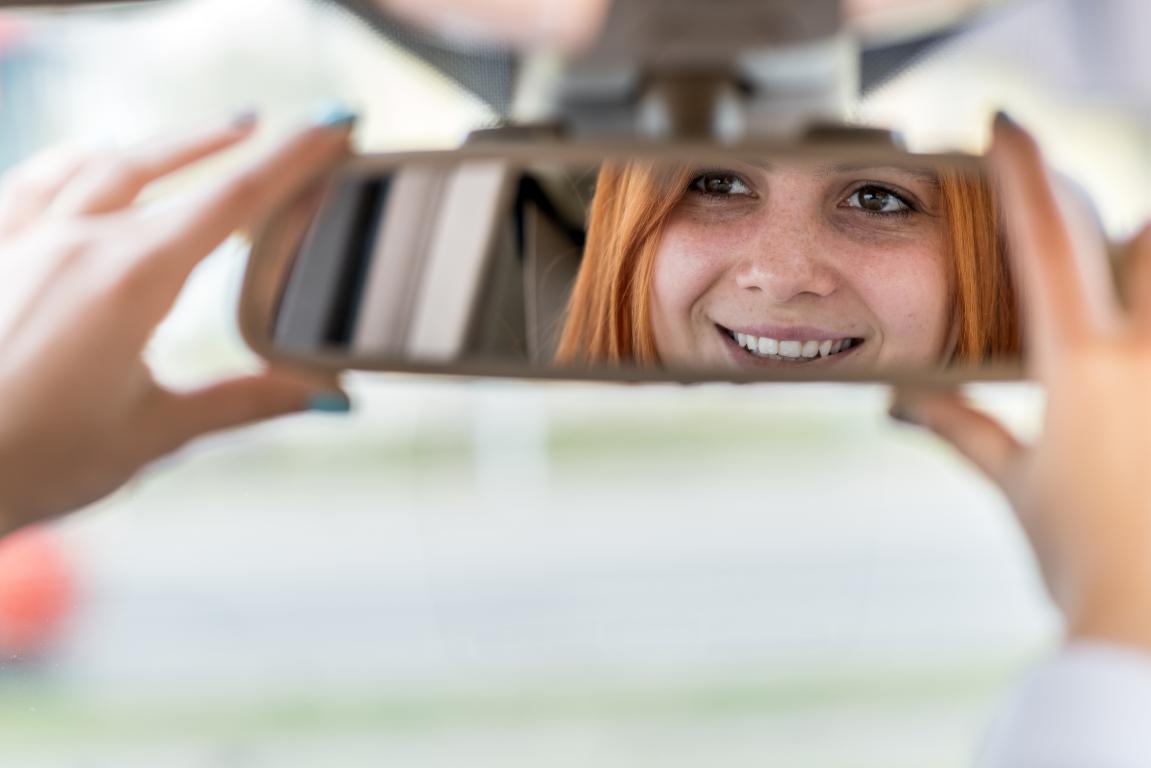 A happy woman with red hair smiles as she adjusts her car's rearview mirror, feeling confident and ready to drive after her recovery.
