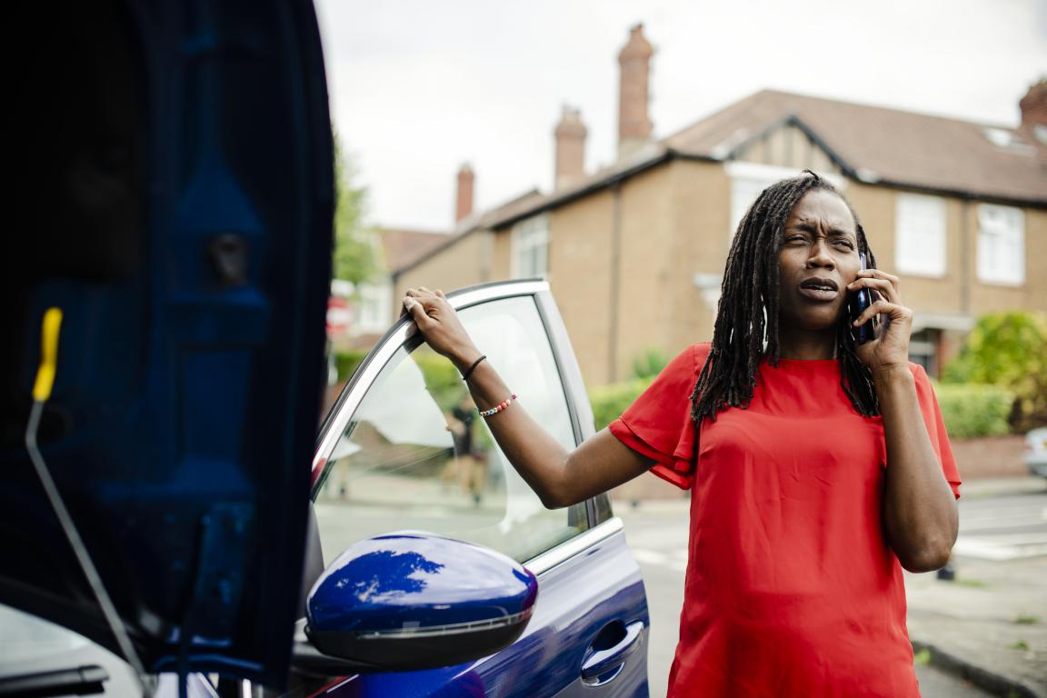 A woman looks stressed on the phone next to her car with the bonnet up, showing the need for accessible medical info in an emergency.
