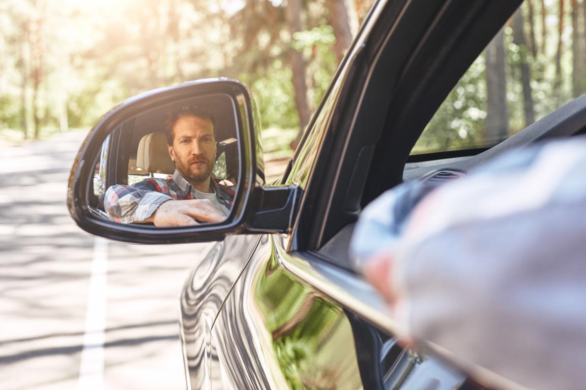 A male driver's face is reflected in the car's wing mirror as he performs a safety check, an important step before driving after surgery.