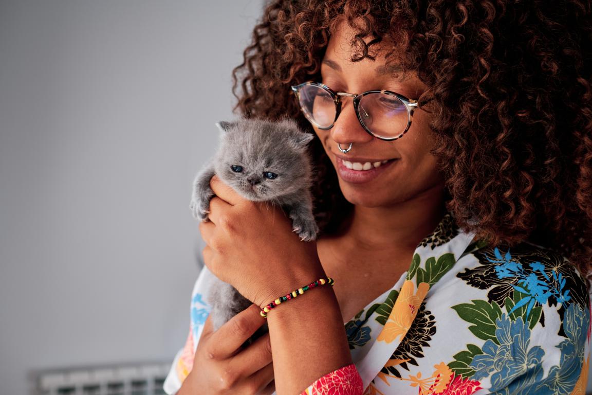 A smiling young woman wearing glasses gently holding a tiny, adorable grey kitten, showing the love and responsibility of a new cat owner.