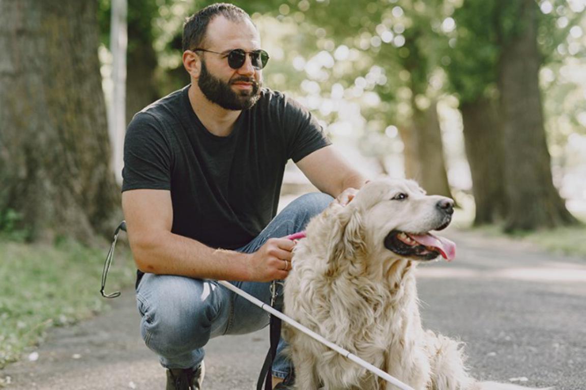 A bearded man with sunglasses and a white cane crouches next to his golden retriever assistance dog on a paved path in a park, both looking relaxed.