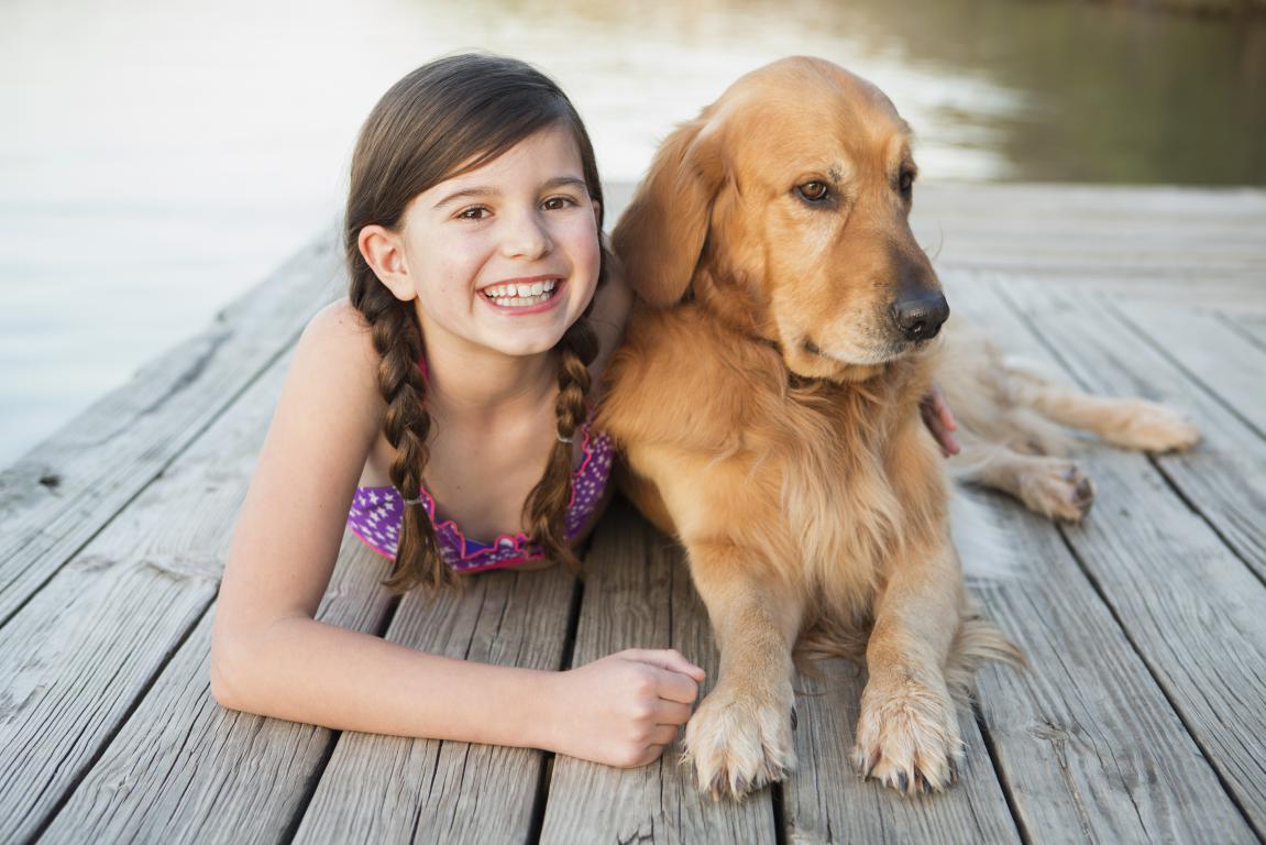 A smiling girl with braids in a swimsuit lies on a wooden dock next to a calm golden retriever, looking towards the camera.