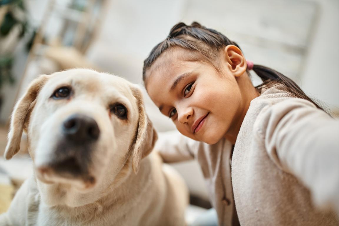 A close-up, low-angle selfie shows a smiling girl with dark hair next to a golden retriever, both looking at the camera.