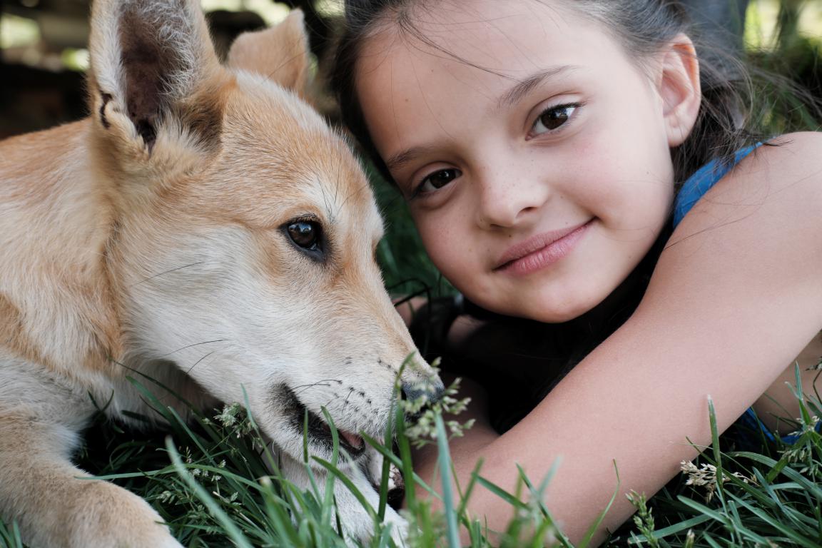 A close-up shot of a girl with dark hair and a light brown dog lying in the grass, both looking gently to the side.