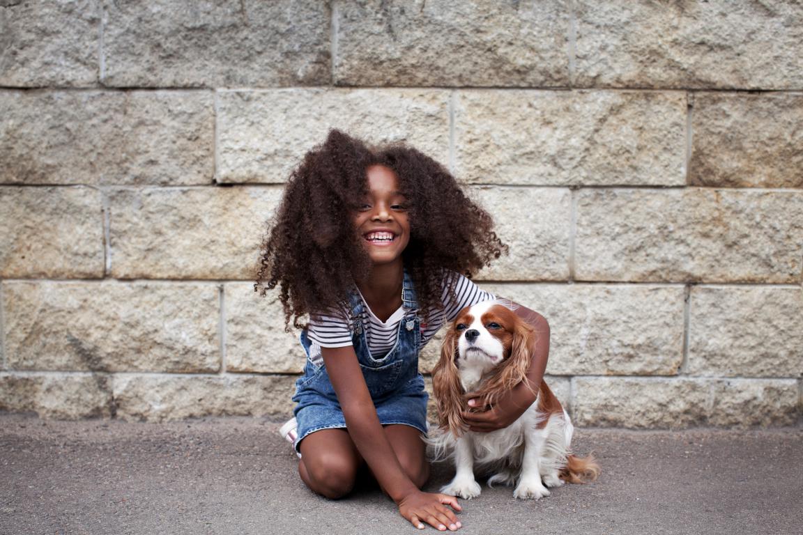  A smiling girl with curly hair, wearing overalls, kneels on pavement next to a Cavalier King Charles Spaniel, both looking towards the camera against a stone wall.