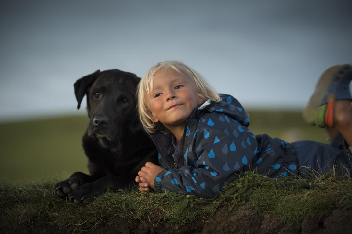 A blonde-haired child in a patterned blue jacket lies on a grassy mound next to a large black dog, both looking towards the camera