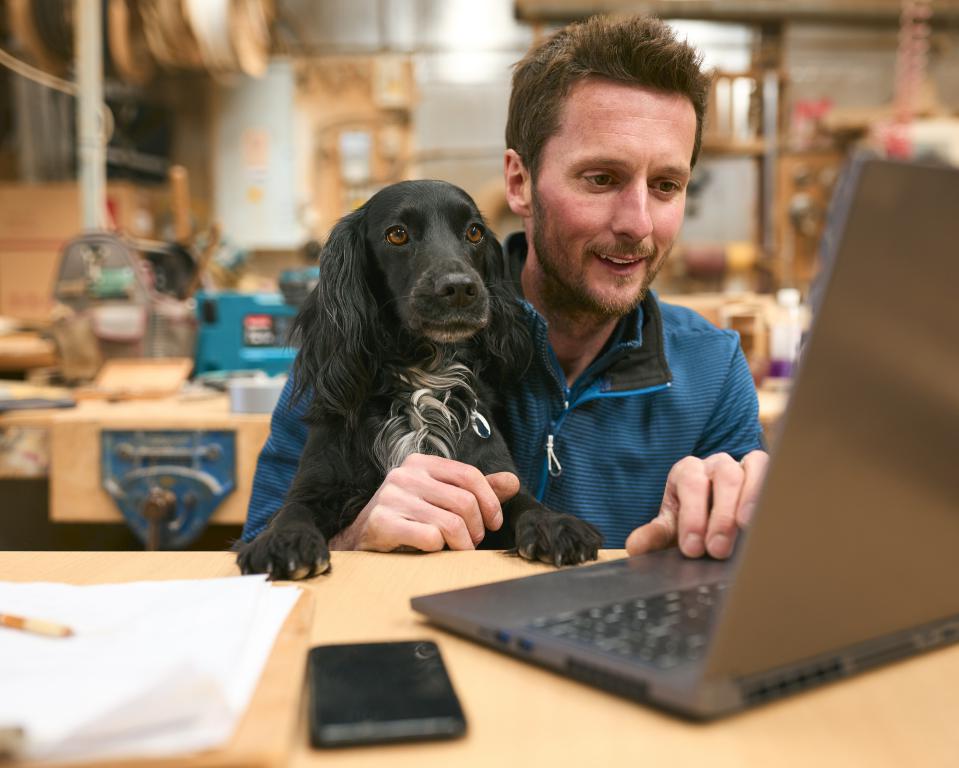 A man with a friendly black spaniel resting its paws on his arms while he works on a laptop at a desk in a workshop