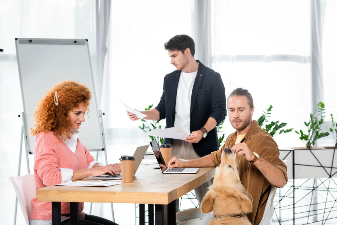 A man at an office desk feeds a treat to a golden retriever assistance dog standing beside him, while a woman works on a laptop nearby.