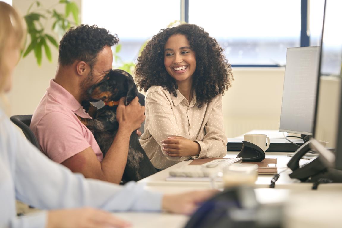 A man happily holds a black and tan dachshund assistance dog while a woman smiles, looking at him, in an open-plan office.