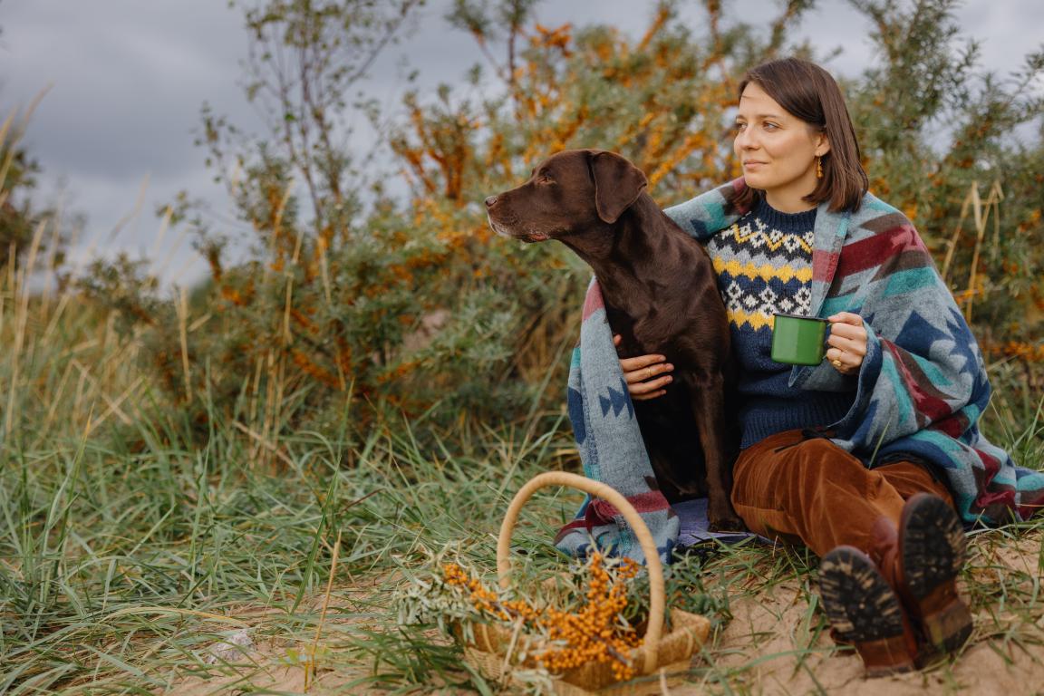 A woman wrapped in a colourful blanket sits on grassy dunes with a thoughtful expression, a brown Labrador-type assistance dog leaning against her. A green mug and a basket of berries are nearby.