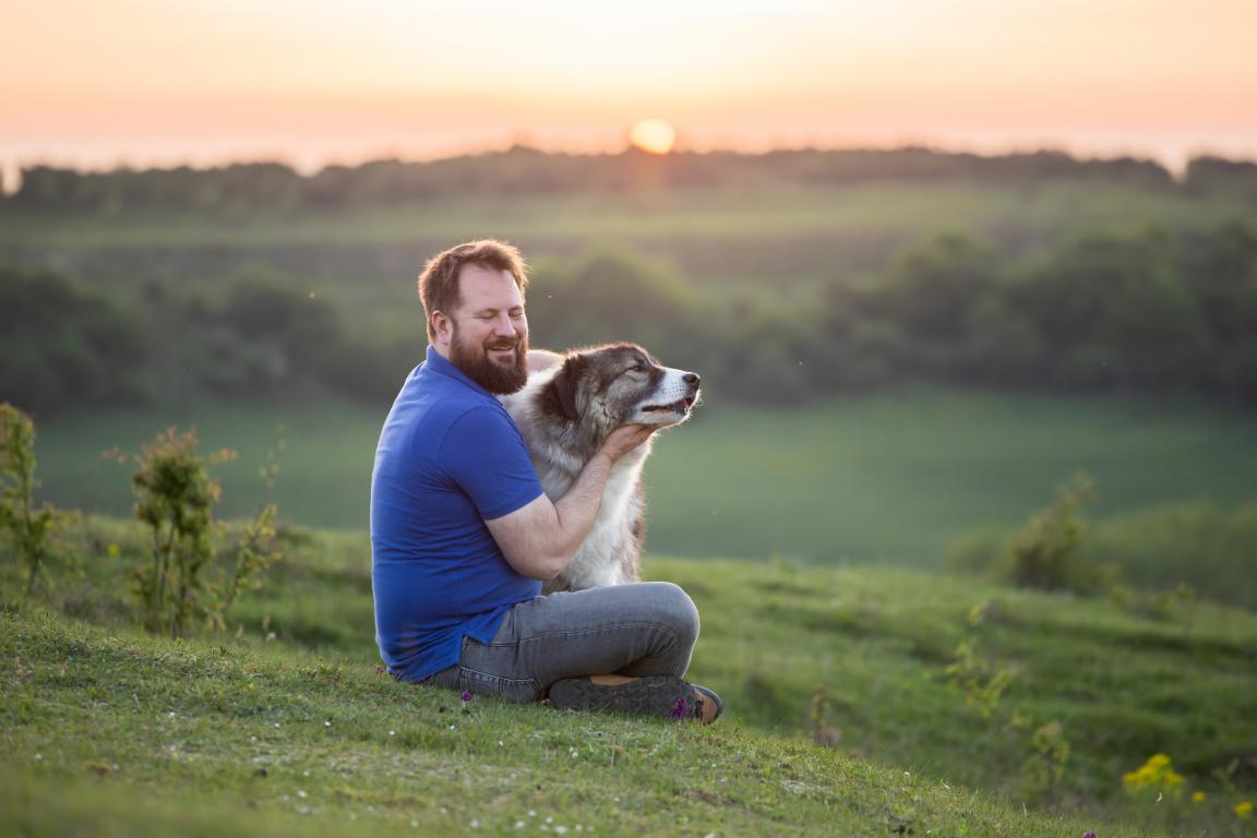 A bearded man in a blue t-shirt and jeans sits cross-legged on a grassy hill at sunset, gently embracing a large fluffy assistance dog with a contented expression