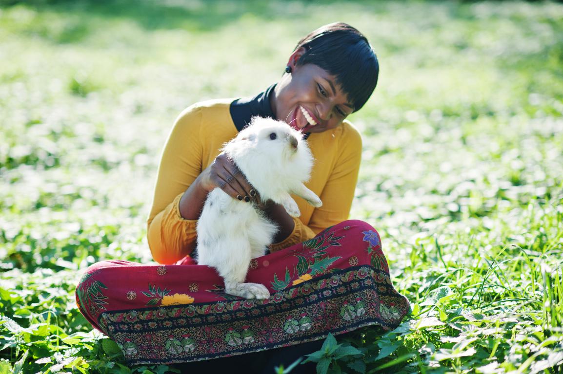 A smiling woman with short dark hair, wearing a yellow top and patterned skirt, sits in a sunny green field, gently holding a fluffy white rabbit.