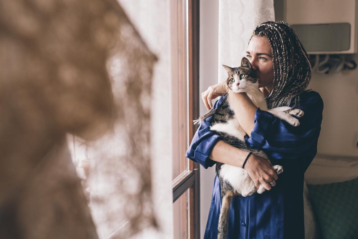 A woman with braided hair, wearing a blue shirt, stands by a window gently hugging a black and white tabby cat, looking out with a calm expression.