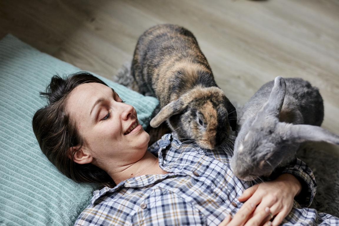 A woman with her eyes closed in contentment lies on her back on a light blue pillow, with two rabbits (one brown and black, one grey) resting on her chest and shoulder.