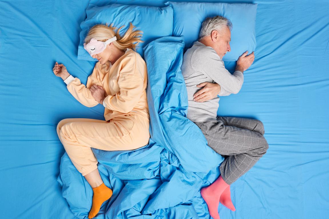  An overhead shot of an older couple lying in separate sleeping positions on a bed with blue sheets. The woman on the left wears a peach-colored loungewear set, an eye mask, and orange socks. The man on the right wears a grey long-sleeved shirt, grey plaid pants, and pink socks. A blue duvet separates them.