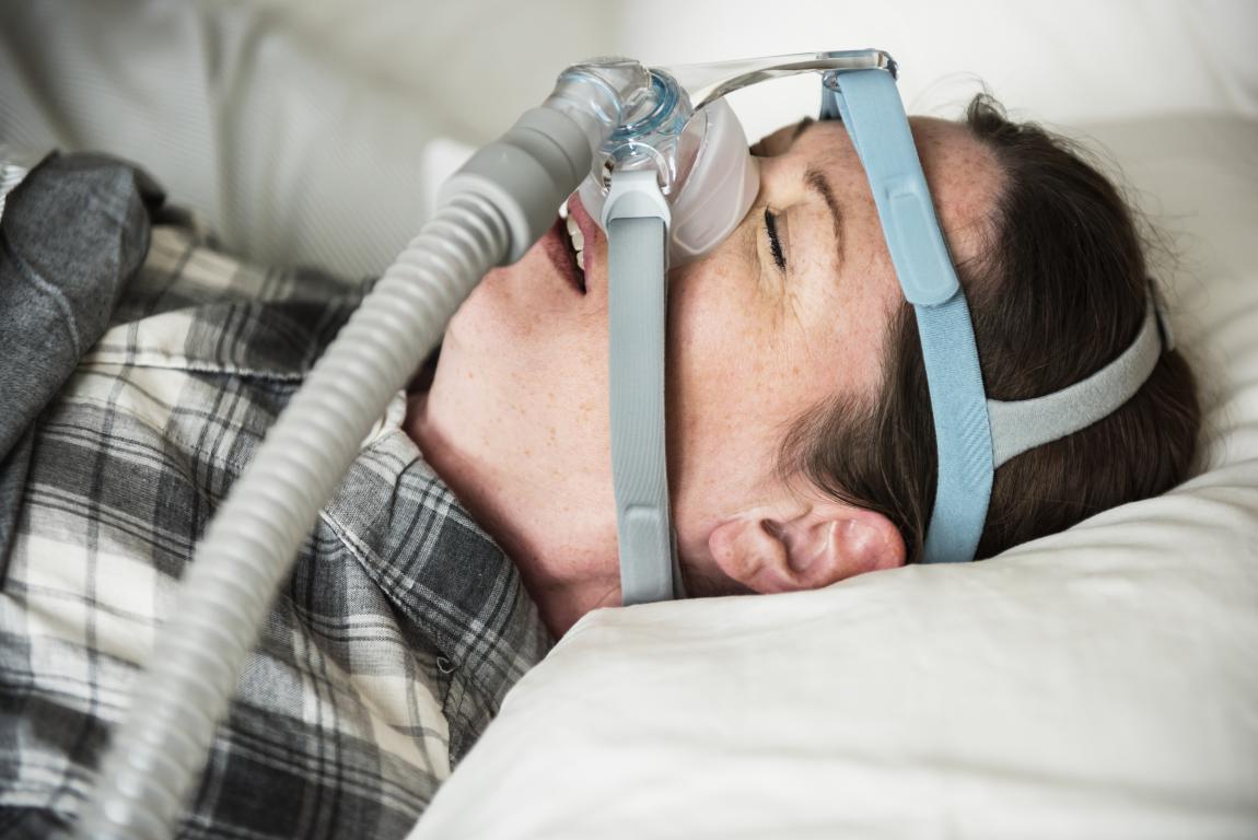 A person with short brown hair and freckles lies on a white pillow, wearing a CPAP mask and hose. They are wearing a grey and white plaid shirt.