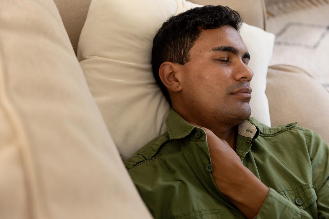 A person with dark hair is lying on a light-colored couch with their eyes closed, appearing to be asleep. They are wearing a green collared shirt and have one hand resting on their chest