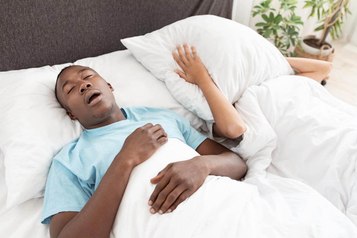 A man with dark skin and short dark hair lies in bed on a white pillow with his mouth open, appearing to be snoring or gasping. He is wearing a light blue t-shirt and is covered by a white duvet. Another person's arm is visible under a pillow next to him.