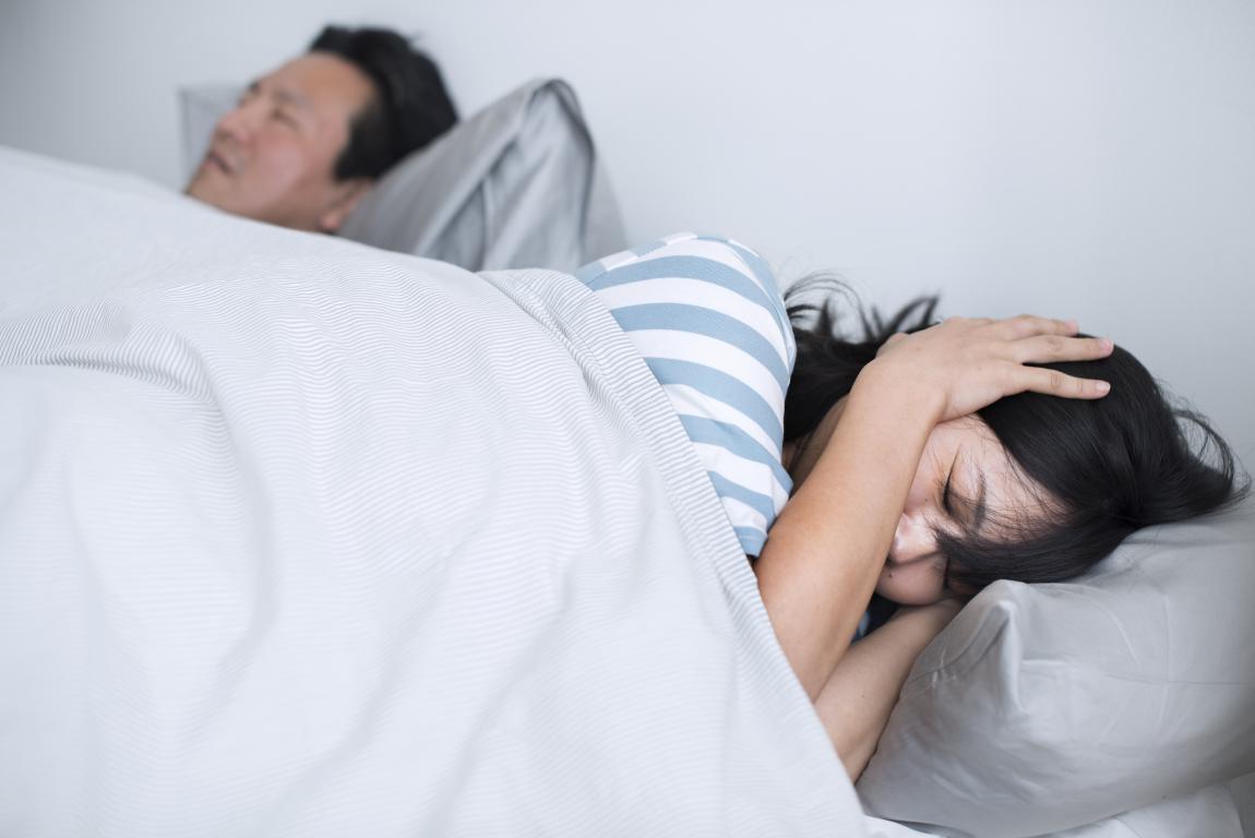 A woman with dark hair lies in bed on a grey pillow, covering her ear with her hand, appearing to be trying to block out noise. She is wearing a blue and white striped shirt and is covered by a light-colored duvet. A man is visible in the background, also lying in bed.