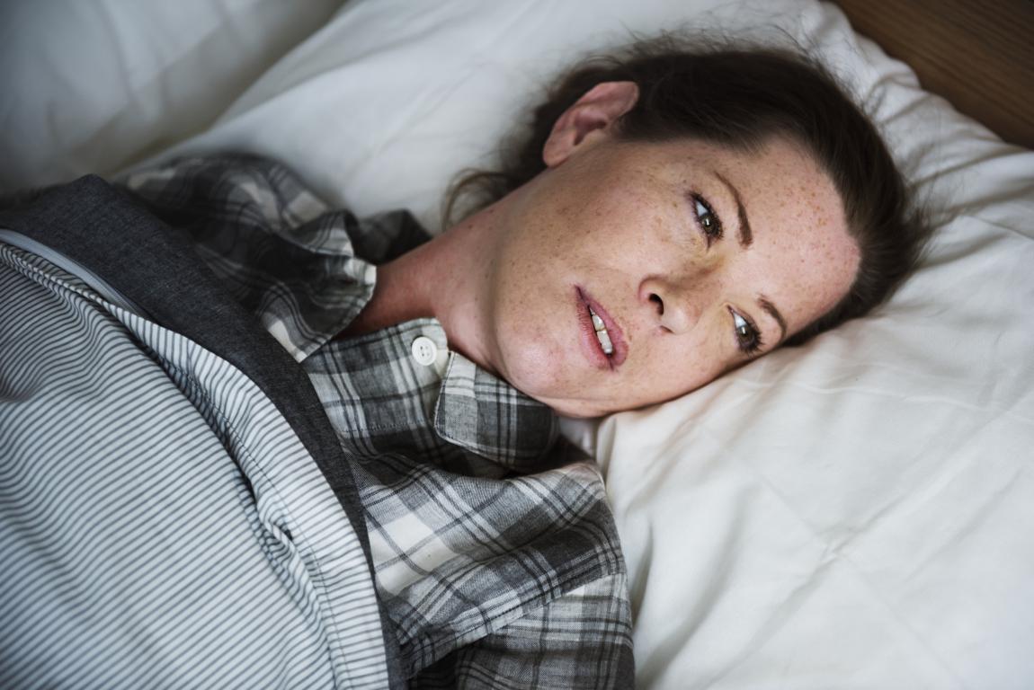 A person with short brown hair and freckles lies in bed on a white pillow, looking to the side with their mouth slightly open. They are wearing a grey and white plaid shirt and are partially covered by a grey and white striped blanket.