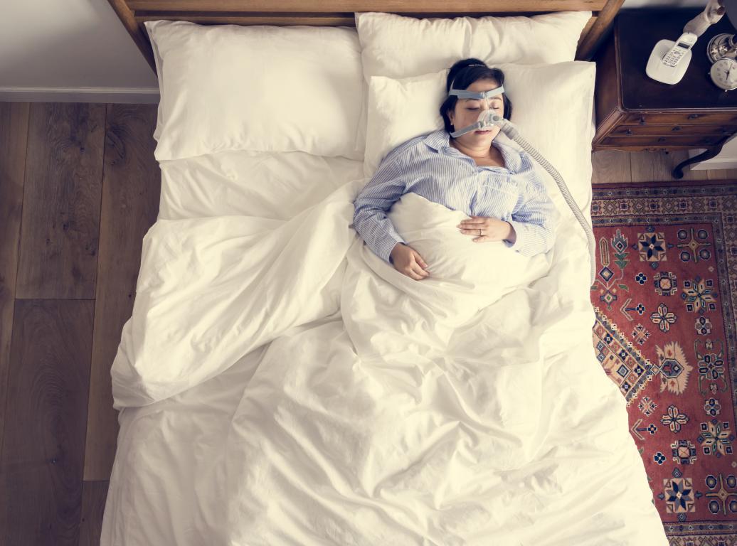  An overhead shot of a person lying in a large white bed, wearing a CPAP mask and hose. They are wearing a blue and white striped shirt and are covered by a white duvet. A nightstand with a phone and alarm clock is visible to the right, next to a patterned rug.