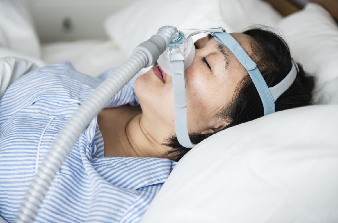 A person with dark hair lies on a white pillow, wearing a CPAP mask and hose. They are wearing a light blue and white striped shirt.