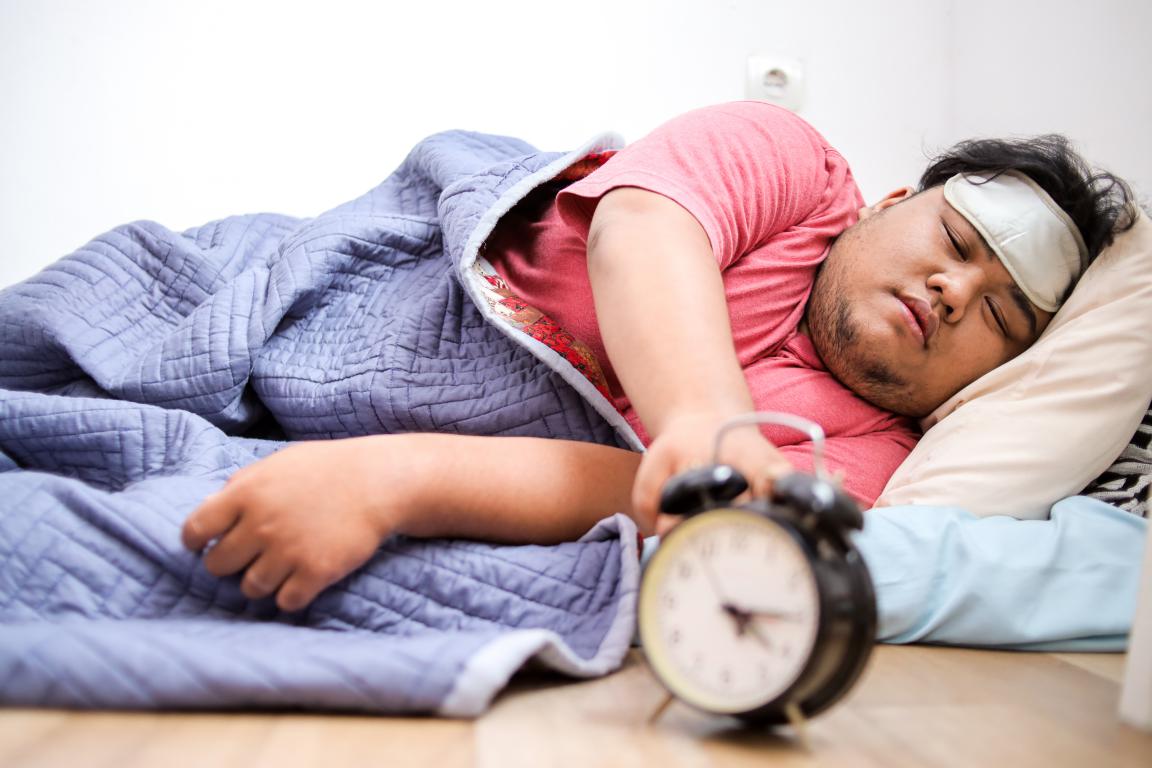 A man in a red shirt and sleep mask reaches for a classic alarm clock on the floor beside his bed, surrounded by blue bedding.