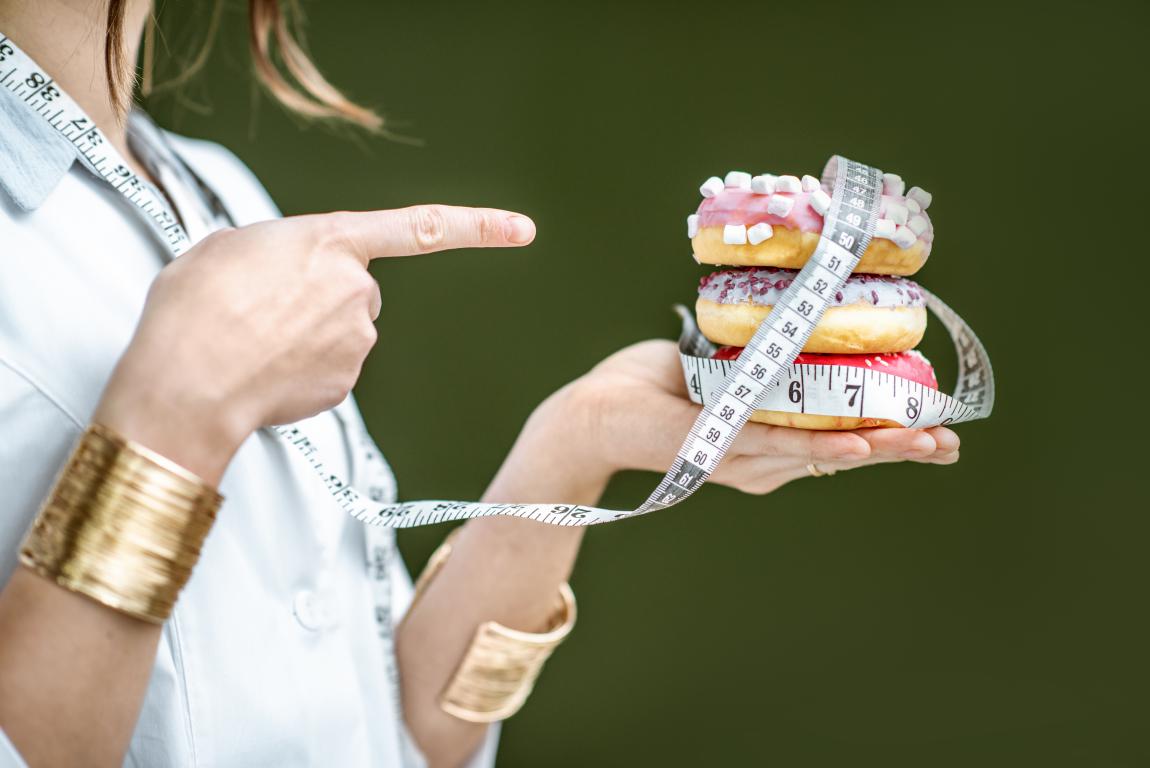A hand holds a stack of colorful frosted donuts wrapped with a measuring tape, while another finger points at them.
