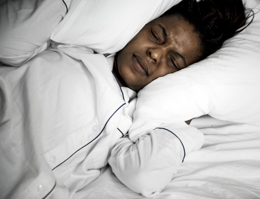 A woman lies in bed, covering her ears with pillows and frowning in discomfort, likely from disturbed sleep.