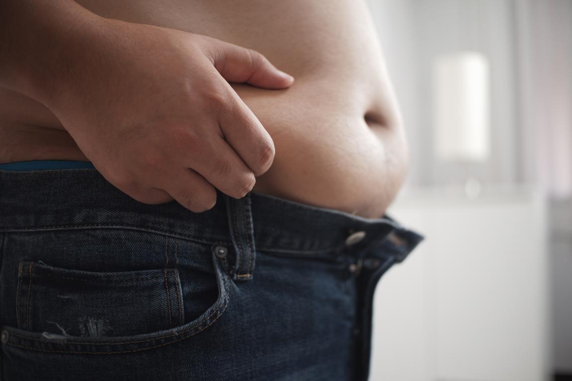 A person's hand pinches excess skin on their stomach above unbuttoned jeans, illustrating abdominal fat.