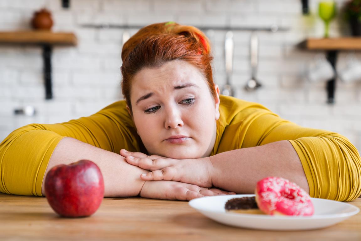 A woman with red hair looks sadly at a plate with a donut and chocolate bar, while a single apple sits untouched nearby on a wooden table.