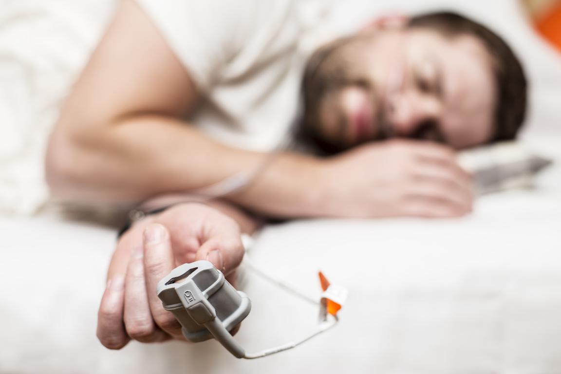 A man sleeps on his side with a pulse oximeter clipped to his finger, connected to a monitoring device in the foreground.