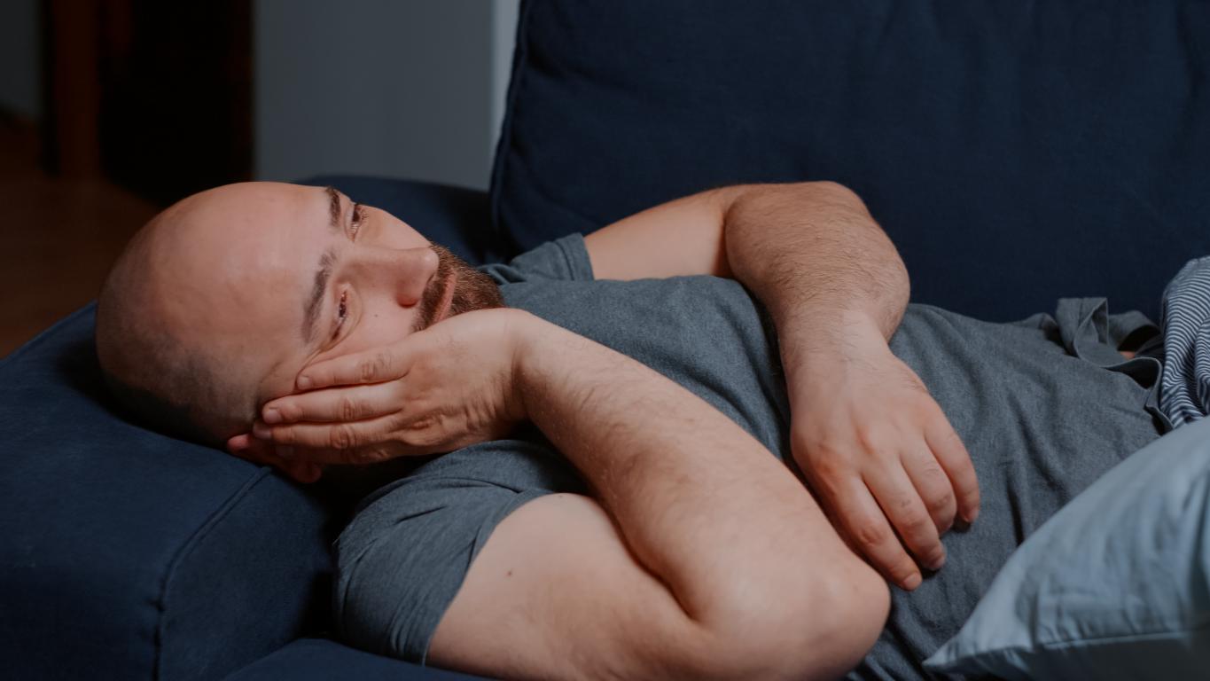 A bald man with a beard lies on a dark blue couch, looking tired and resting his head on his hand.