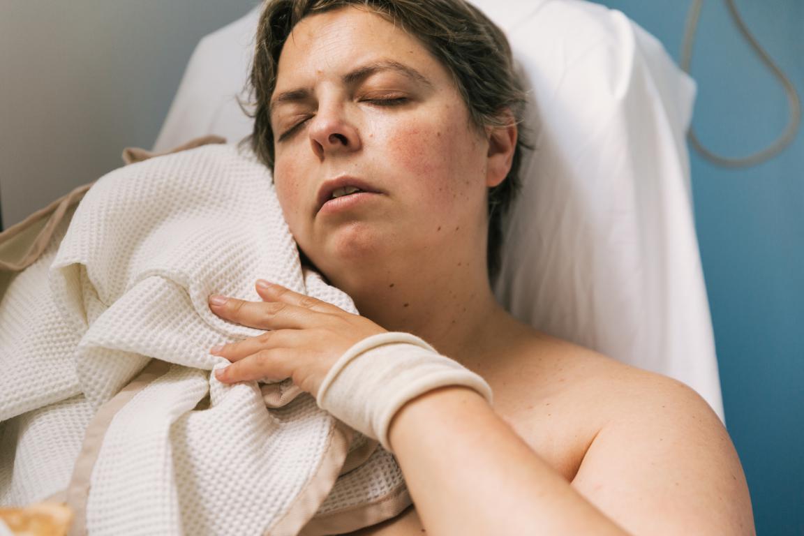 A woman with her eyes closed lies in a bed or medical chair, covered by a white blanket, with a white wristband on her arm.