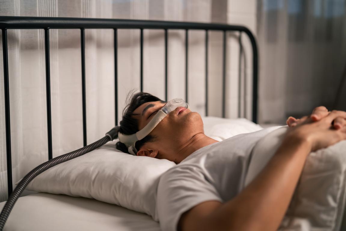 A man lies on his back in bed, sleeping with a CPAP mask over his nose, connected to a machine