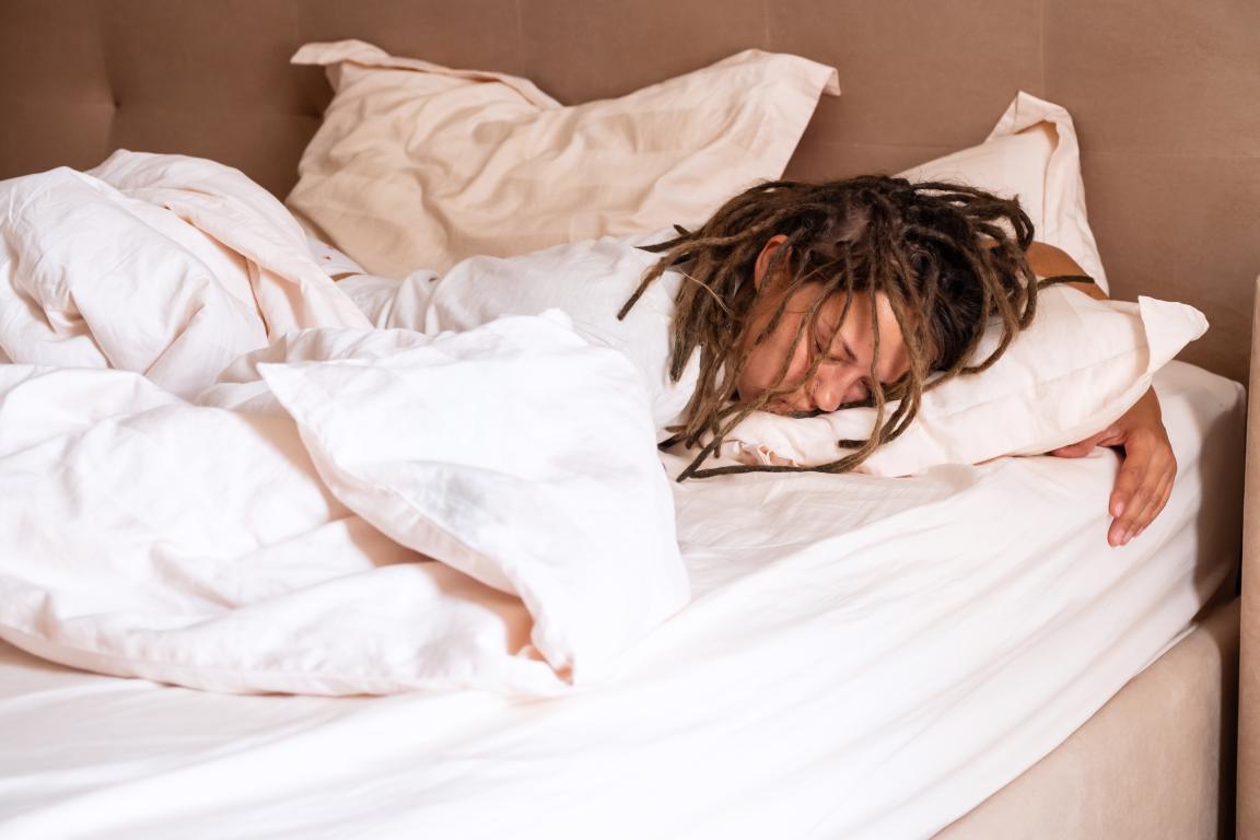 A woman with dreadlocks sleeps on her side, face buried in a pillow, covered by a white duvet.