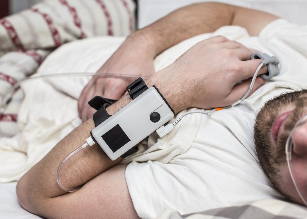 A close-up of a man's arm and chest as he lies in bed, wearing a portable sleep study device on his wrist with sensors on his fingers.