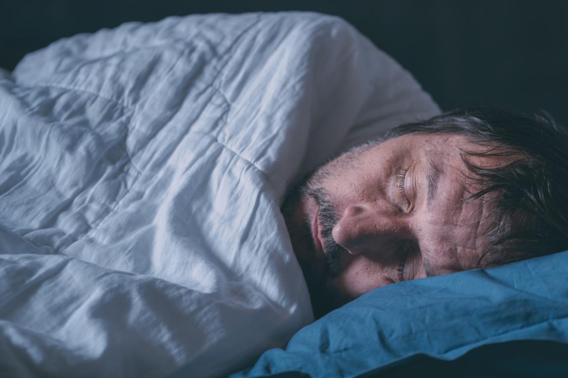 A man with a beard sleeps peacefully under a white duvet, with his head on a blue pillow, in a dimly lit room.