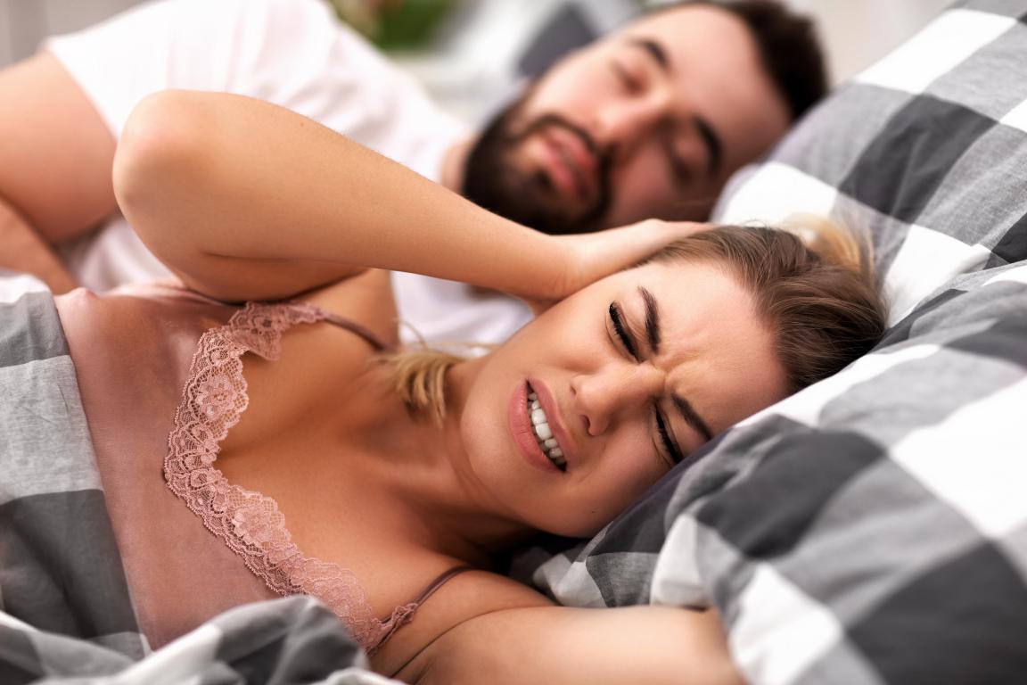  A woman in a pink camisole lies in bed with her hands pressed over her ears, wincing in frustration. A man is partially visible asleep behind her, indicating snoring