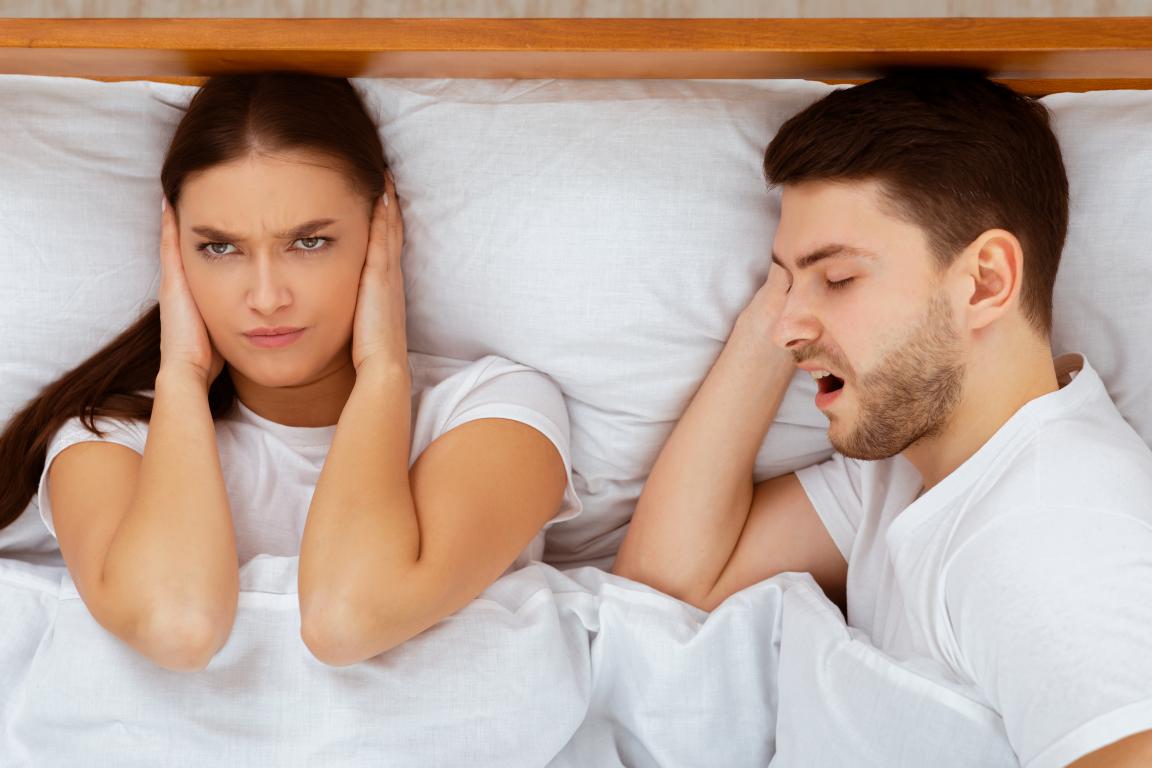  A woman with long dark hair lies in bed, covering her ears with her hands and looking annoyed. Next to her, a man lies on his back with his mouth open, clearly snoring.
