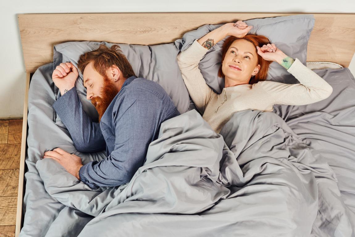 A man with a beard sleeps peacefully on his side under a grey duvet. Beside him, a woman with red hair stretches happily, looking refreshed after a good night's sleep.