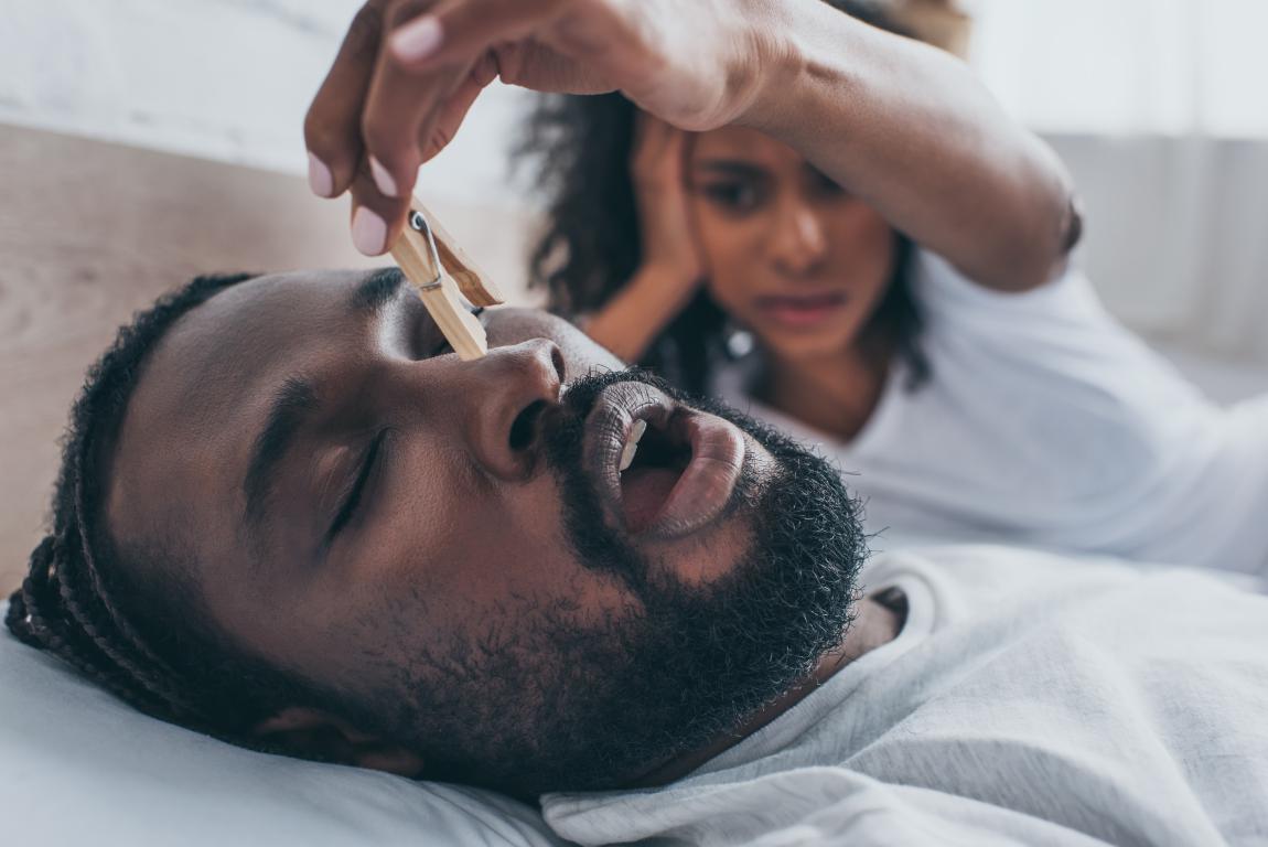  A close-up of a Black man with a beard lying on his back, mouth open, as a hand holds a clothespeg near his nose, looking frustrated