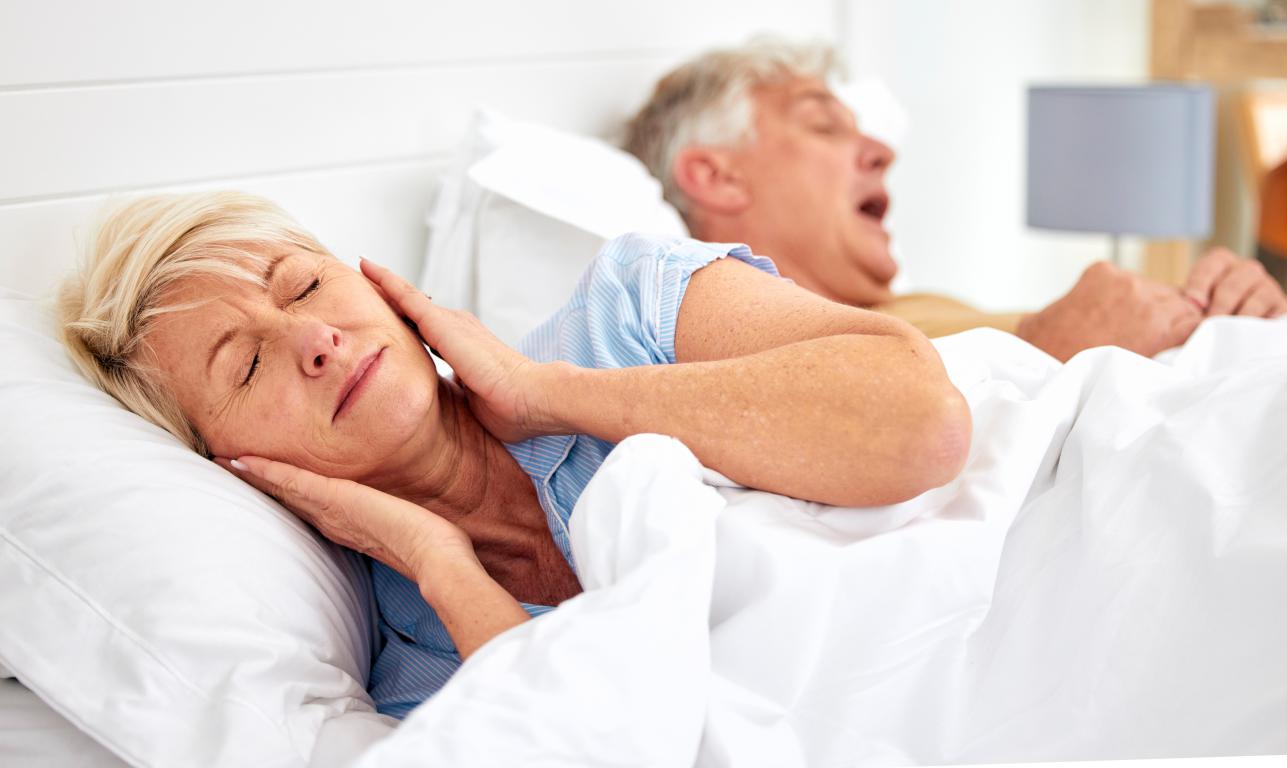 An older woman with blonde hair lies in bed, hands pressed over her ears with a pained expression, trying to block out the sound of a man snoring loudly in the background